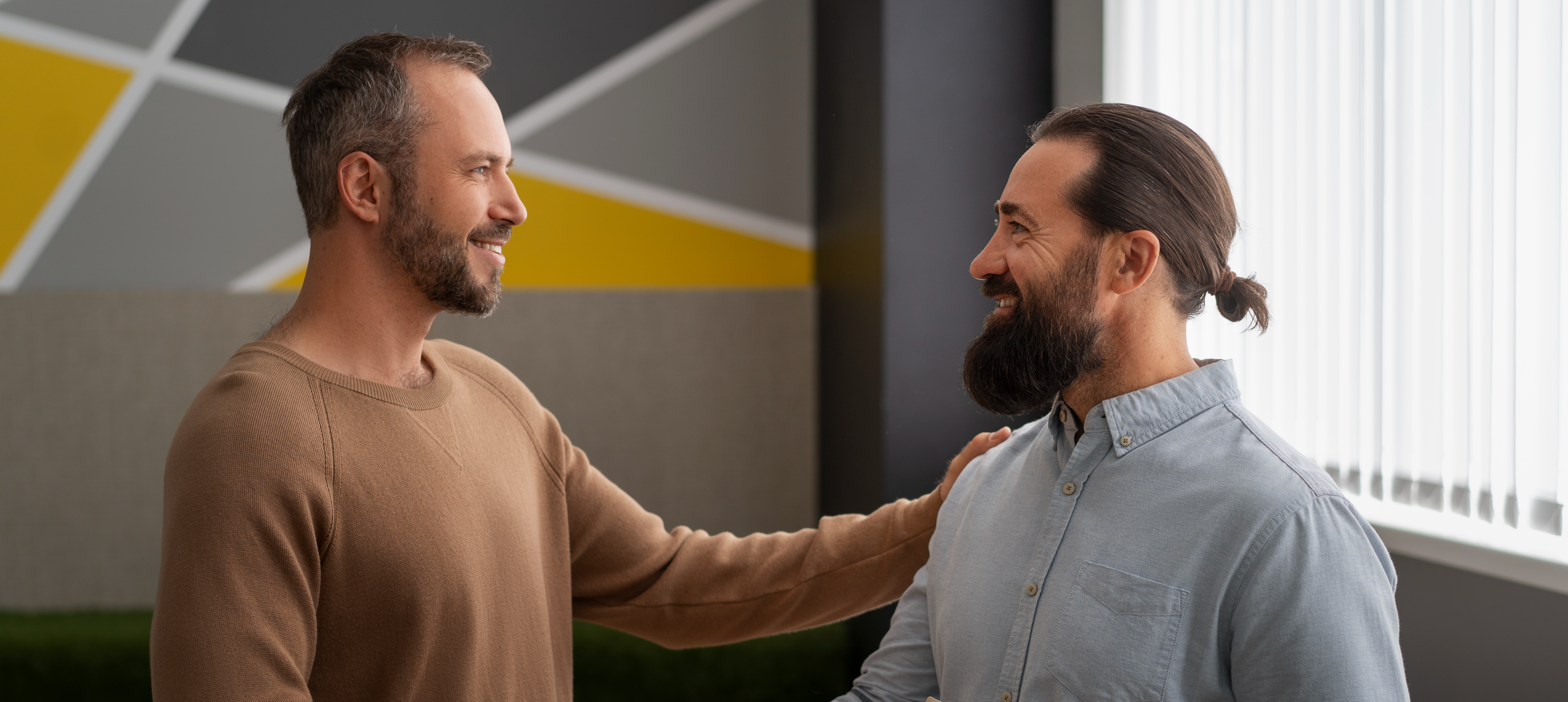 Dois homens em ambiente corporativo cumprimentam-se com um aperto de mãos, demonstrando parceria e cordialidade. Um deles segura uma pasta com documentos.