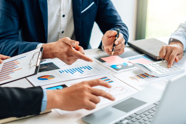 Equipe de negócios analisando gráficos e dados financeiros sobre a mesa, com uso de calculadora, laptop e prancheta durante reunião de planejamento estratégico.